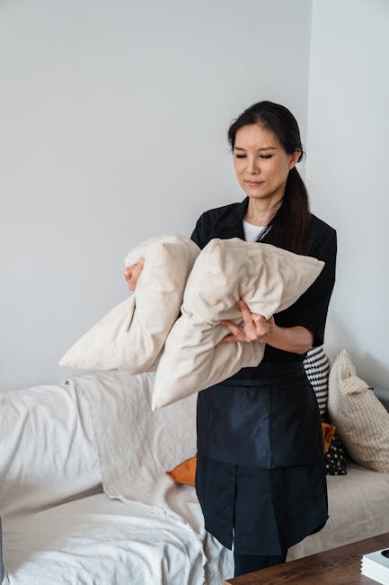 A woman dressed in black scrubs holding two beige pillow covers while standing in a living room with a white sofa that has a cream-colored cushion and an orange cushion. The room features a white wall