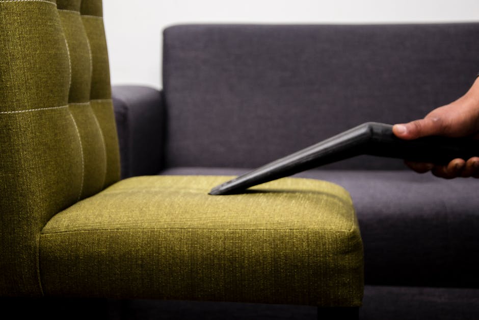 A woman dressed in black scrubs holding two beige pillow covers while standing in a living room with a white sofa that has a cream-colored cushion and an orange cushion. The room features a white wall